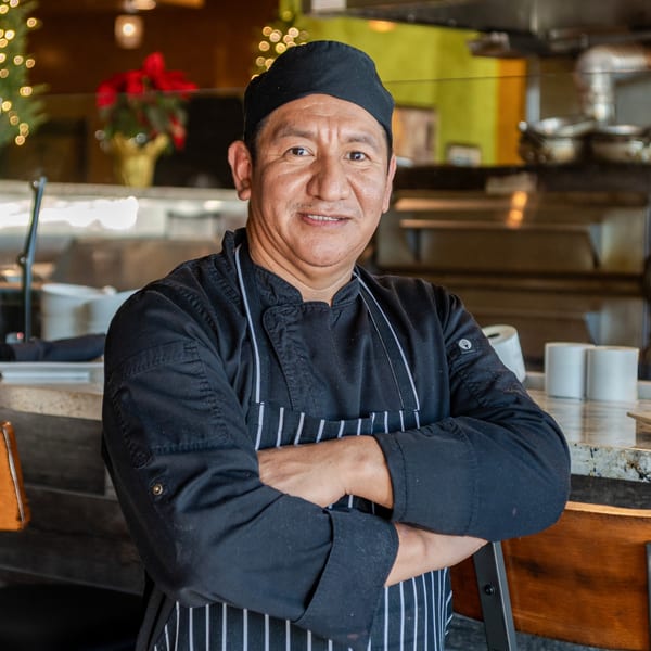 Smiling Mona Lisa Cucina chef in black uniform and striped apron with arms crossed.