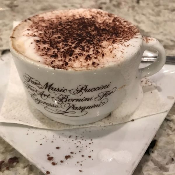 A white cup of cappuccino with frothy milk and cocoa powder, on a white saucer.