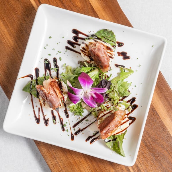 Italian appetizer with grilled meat, mozzarella, greens, balsamic glaze, and an orchid on a white plate.