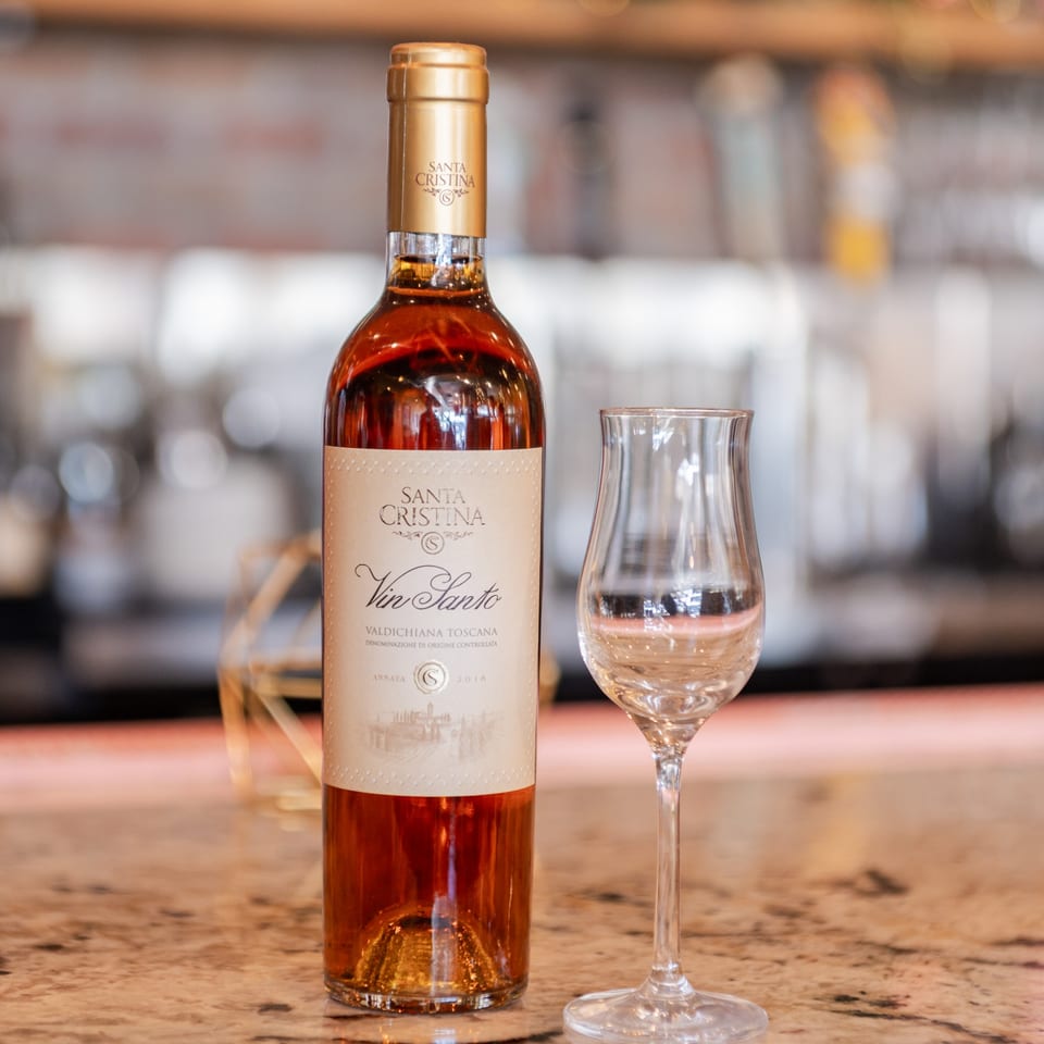A bottle of Santa Cristina Vin Santo dessert wine next to an empty tasting glass on a counter.