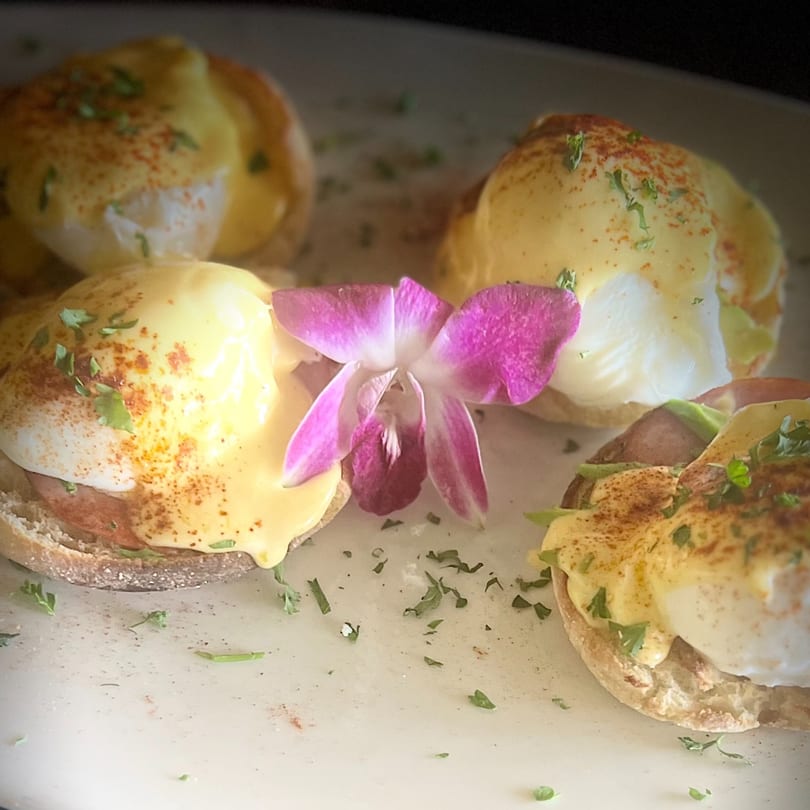 Four eggs Benedict with hollandaise sauce, paprika, and parsley, garnished with a purple orchid.
