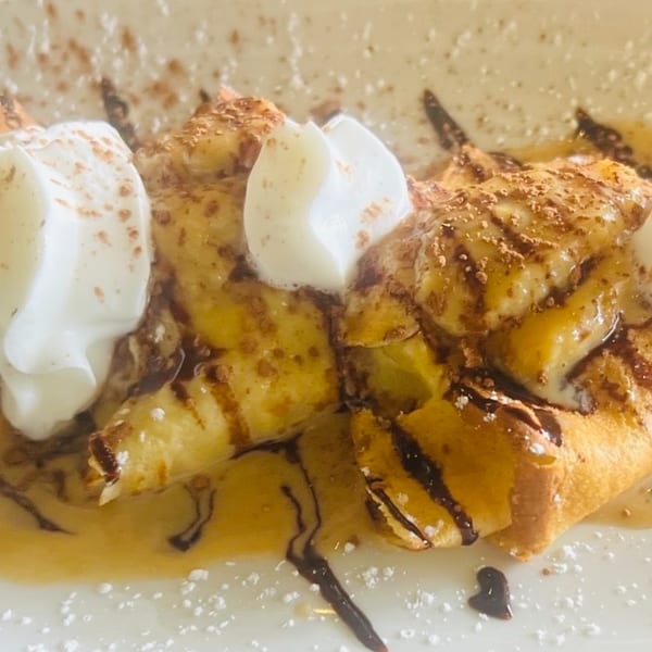 Close-up of golden crepes topped with whipped cream, chocolate drizzle, and powdered sugar.
