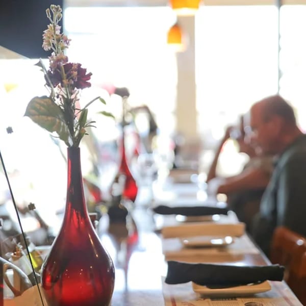 A red vase with flowers on a table, with blurred diners and bright windows in the background.