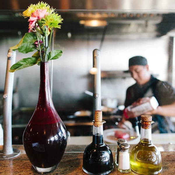 A chef cooks in the background, with a vase of flowers, olive oil, and balsamic vinegar in the foreground.