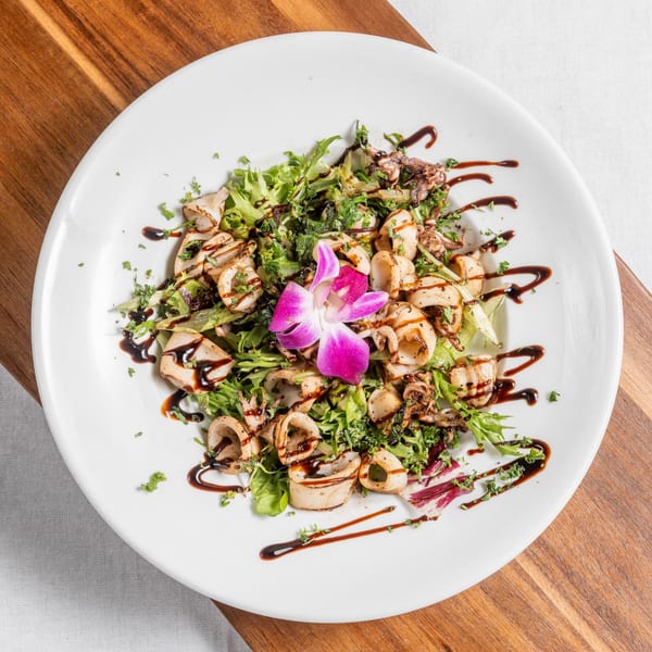 Grilled calamari salad with mixed greens, balsamic glaze, and a pink orchid garnish on a white plate.
