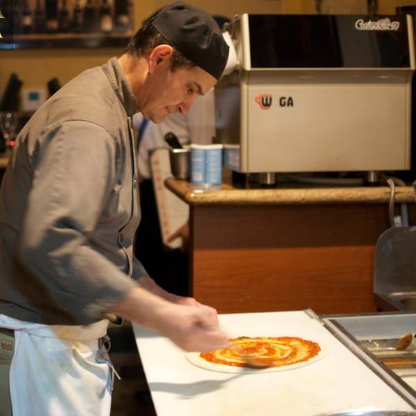 A chef in a black cap and grey shirt spreads tomato sauce on a pizza base.