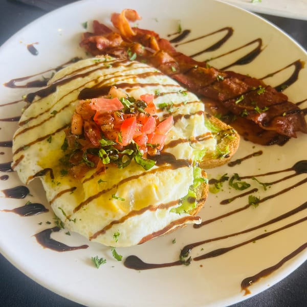 Avocado toast with fried eggs, diced tomatoes, herbs, and bacon, drizzled with balsamic glaze.