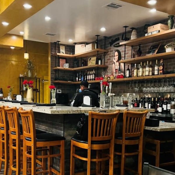 A person sits at the Mona Lisa Cucina bar with wooden stools, shelves of wine, and a Mona Lisa portrait.