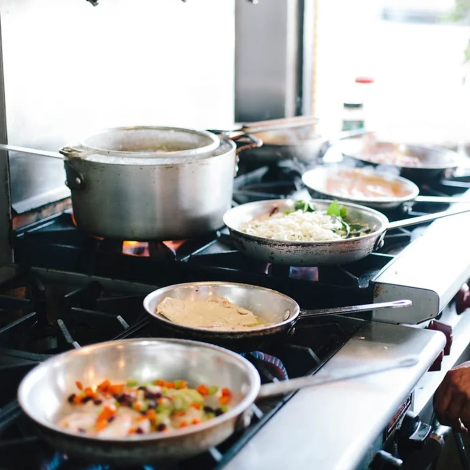 Multiple pans and a pot cooking on a commercial stove, preparing Italian dishes.