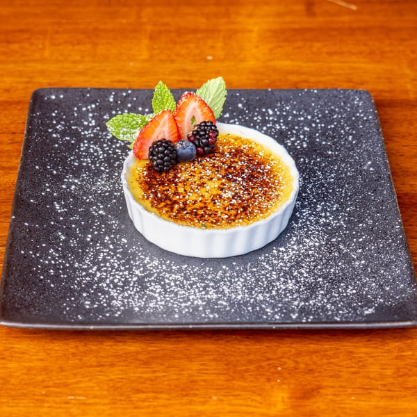 Crème brûlée dessert topped with fresh strawberries, blackberries, blueberries, and mint on a dark plate.