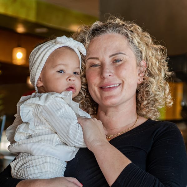 A smiling woman with curly hair holds a baby wearing a white ruffled outfit and headband.