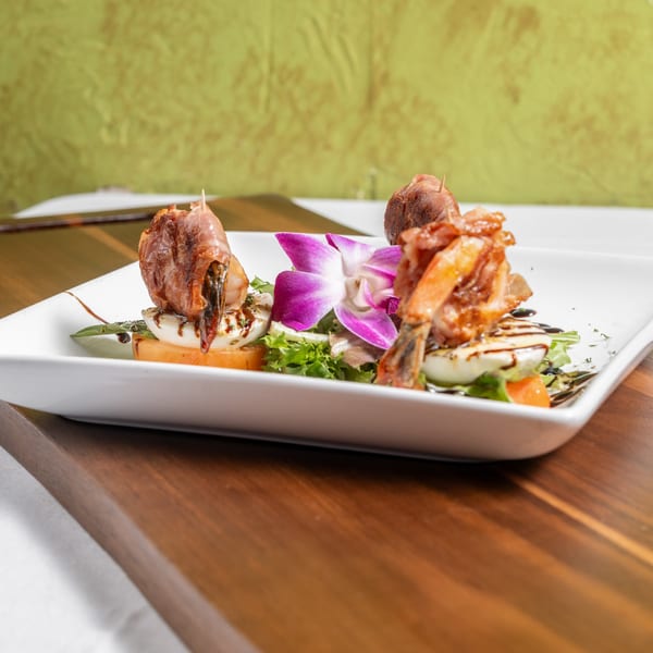 Italian appetizer with prosciutto-wrapped shrimp, mozzarella, tomatoes, greens, and a purple orchid.