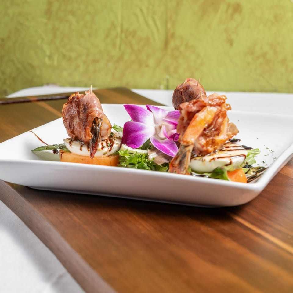 Italian appetizer with prosciutto-wrapped shrimp, mozzarella, tomatoes, greens, and a purple orchid.